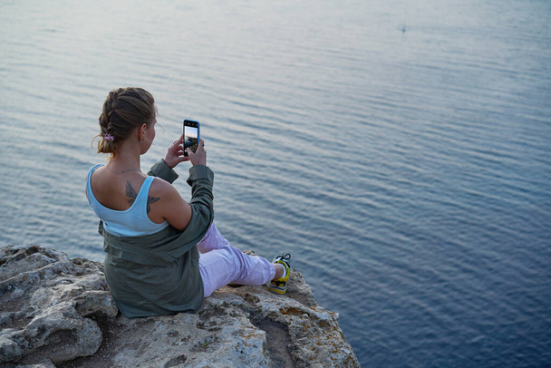 Frau sitzt auf einem Felsen, blickt auf die Meereslandschaft, den Sonnenuntergang, macht Fotos und genießt die Aussicht und die frische Luft. - Foto, Bild