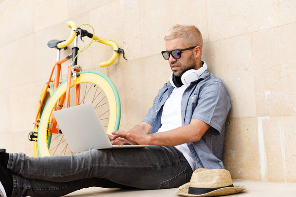 Ισπανόφωνος νομάδας ελεύθερος επαγγελματίας εργάζεται στο laptop του σε εξωτερικούς χώρους δίπλα σε ένα αστικό πολύχρωμο ποδήλατο. - Φωτογραφία, εικόνα