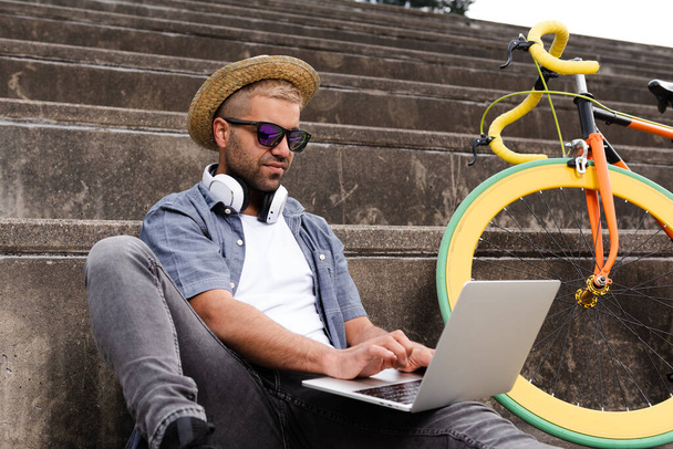 Latynoski hipsterski wolny strzelec pracujący na laptopie na świeżym powietrzu obok kolorowego roweru. - Zdjęcie, obraz