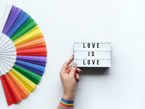 LGBTQ pride month decor. Text Love is Love on lightbox. Rainbow fan on white background. Human hand with rainbow nail varnish holds light box. June, LGBT pride month pride simple minimal decor. - Foto, imagen