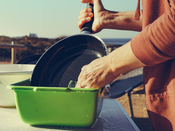 Mature woman washing up dishes in bowl on fresh air. Dishwashing outdoor on camping site, sea shore - Foto, Imagem