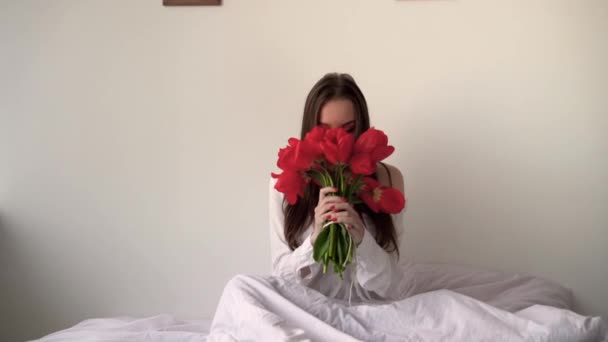 Una signora con un mazzo di fiori si siede sul letto in camera da letto, annusando tulipani e starnuti allergie. Mattina e millenario su biancheria da letto e un materasso ortopedico. Il concetto di moglie del mattino. - Filmati, video