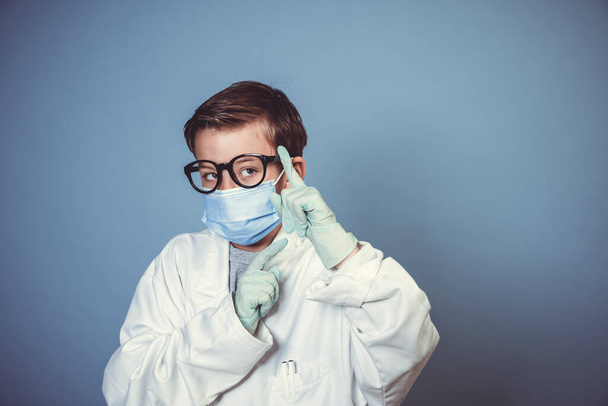 cool boy with mouth guard, black glasses and rubber gloves with white lab coat or doctor coat on blue background in the studio - Фото, изображение