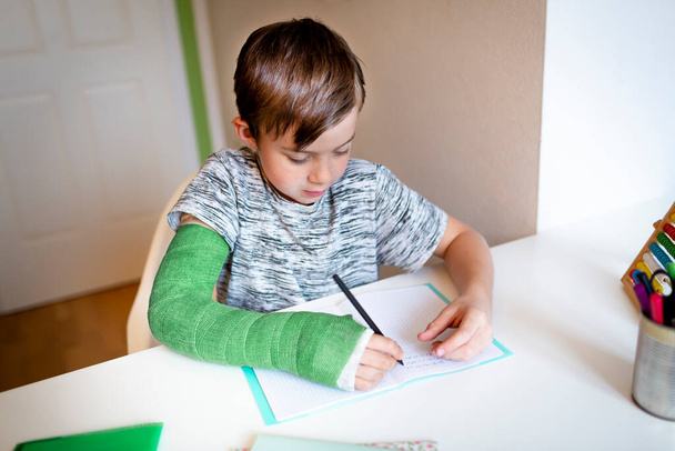 cool boy with green arm cast is sitting in his room and is writing something and is doing housework - Photo, Image