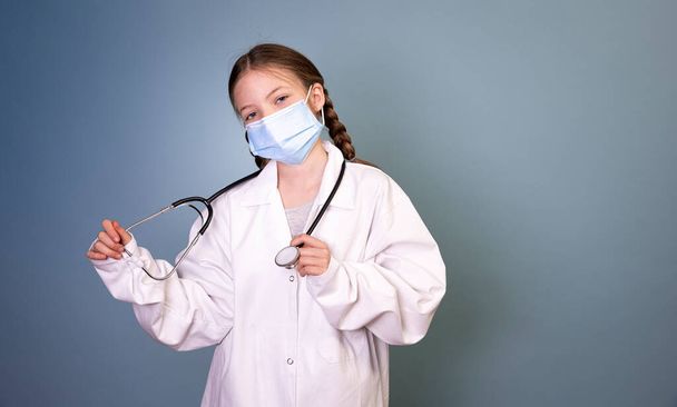 pretty young girl with 2 braids dressed as a doctor with protective mask and stethoscope in front of blue background - Фото, изображение