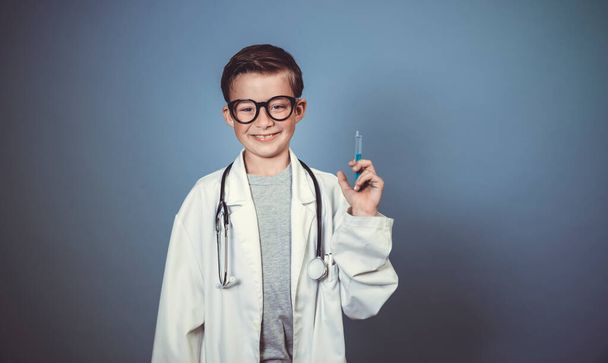 cool young boy is disguised as a doctor with a white doctor smock and plays with syringe and stethoscope in front of blue background - Fotoğraf, Görsel
