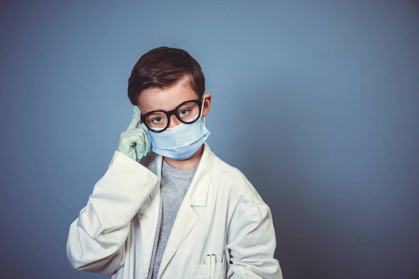 cool boy with mouth guard, black glasses and rubber gloves with white lab coat or doctor coat on blue background in the studio - Foto, Imagem