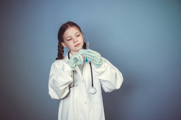 cool young girl dressed as female doctor with stethoscope and syringe and rubber gloves posing in front of blue background - Фото, изображение