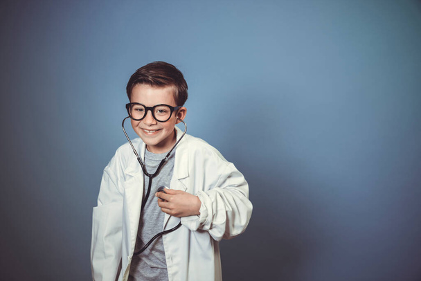 cool young boy is disguised as a doctor with a white doctor smock and plays with syringe and stethoscope in front of blue background - Foto, afbeelding