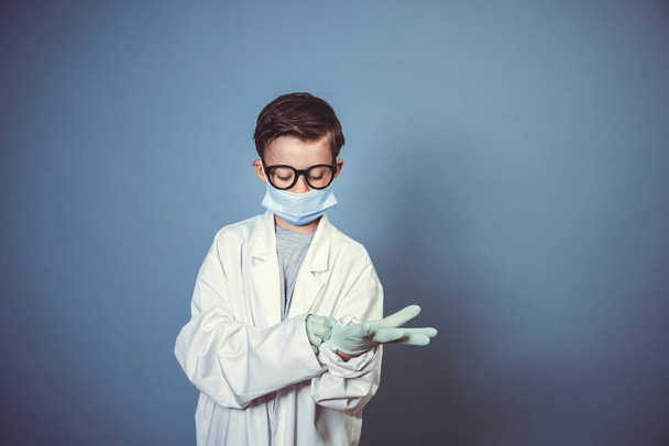 cool boy with mouth guard, black glasses and rubber gloves with white lab coat or doctor coat on blue background in the studio - Foto, immagini