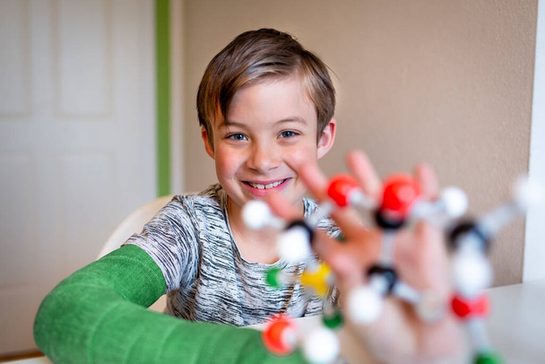 cool schoolboy with green arm plaster cast sits in his room and plays with model of molecule - Foto, Imagem