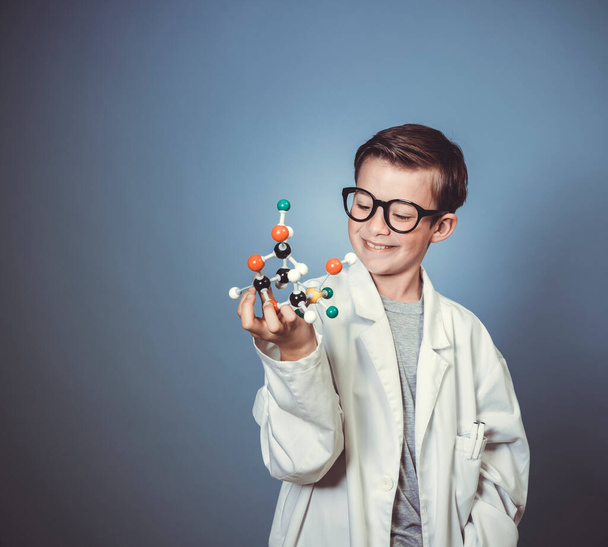 cool young boy is dressed as scientist with white lab coat and holding model of molecules in hand and wearing black glasses - Photo, Image