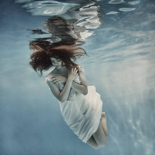              A girl with long dark hair in a white dress with glitters floats underwater as if floating in zero gravity on a light background                   - Photo, image