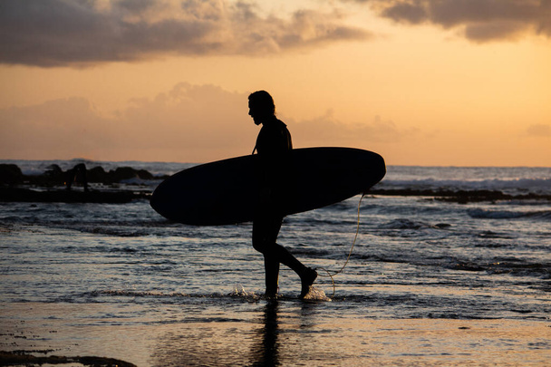 silueta de un surfista que sale del agua al atardecer con su tabla de surf. - Foto, imagen