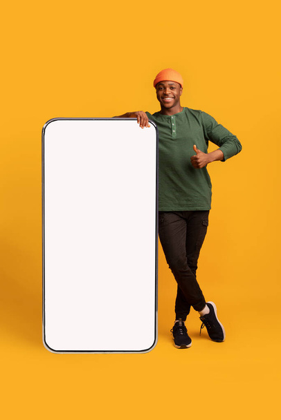 Щасливий чорний хіпстер хлопець спирається на великий порожній смартфон, жестикулюючи великий палець вгору
 - Фото, зображення