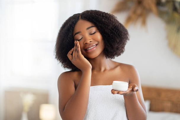 Beautiful young black woman applying face cream from jar, wearing bath towel, enjoying spa procedure at home - Foto, imagen