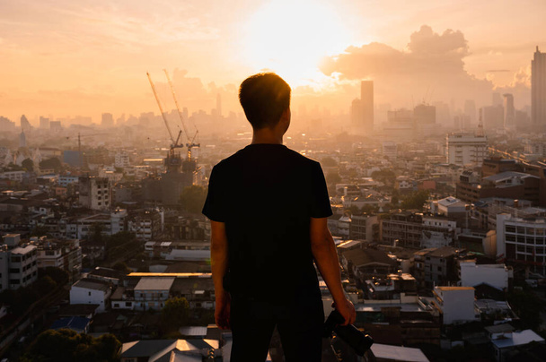 Jovem asiático homem com câmera em pé no topo do edifício e vista do nascer do sol na cidade de Bangkok, na Tailândia - Foto, Imagem
