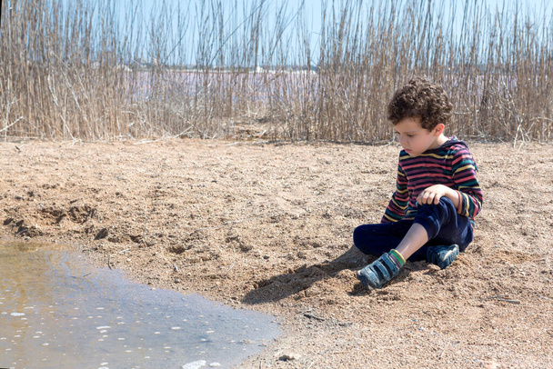 4-5 yaşlarında, beyaz saçlı, güneşli bir günde gölün kenarında kumla oynayan, renkli çizgili gömlekli bir çocuk. - Fotoğraf, Görsel