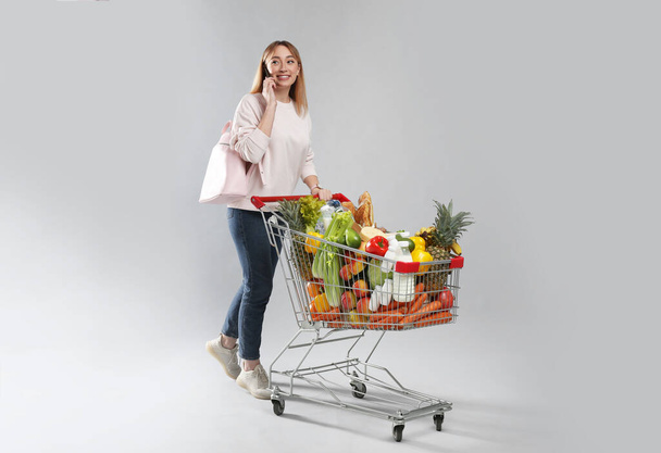 Young woman with shopping cart full of groceries talking on phone against grey background - 写真・画像