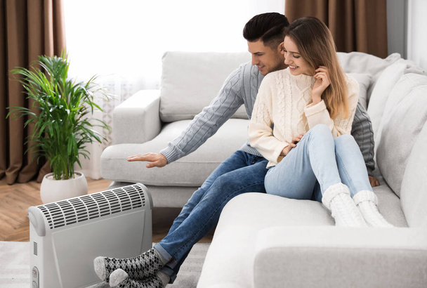 Happy couple sitting on sofa near electric heater at home - Fotografie, Obrázek