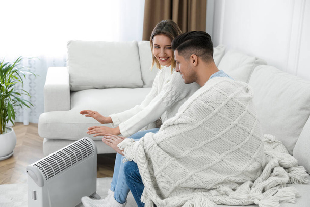 Happy couple sitting on sofa near electric heater at home - Zdjęcie, obraz