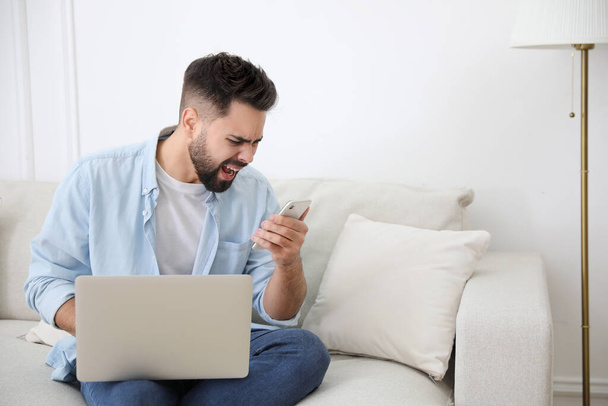 Emotional young man with laptop and smartphone at home. Online hate concept - Foto, Imagem