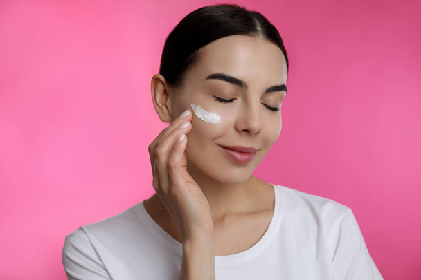 Young woman applying facial cream on pink background - Fotó, kép