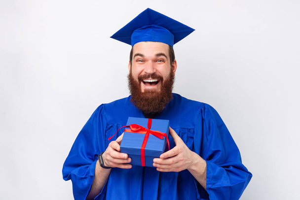 Amazed young bearded man wearing blue bachelor and holding a gift box. - Photo, Image
