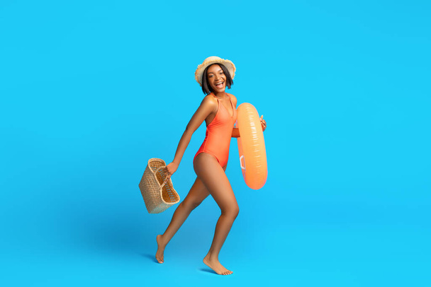 Swimming is fun. Black woman in swimsuit going to pool with inflatable ring and straw bag, blue background, copy space - Photo, Image