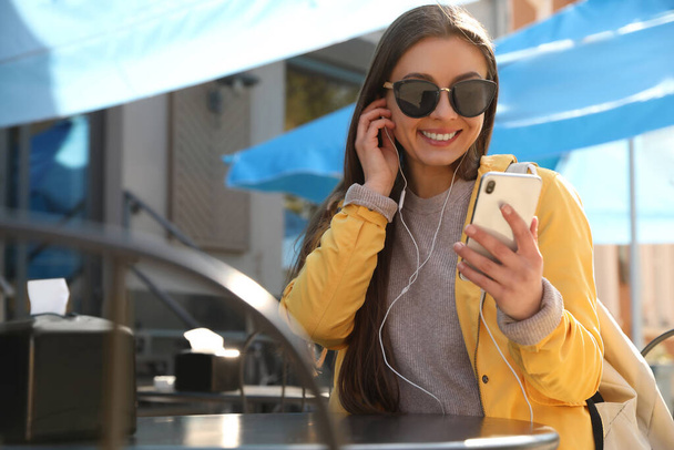 Mujer joven feliz con auriculares y teléfono móvil escuchando música en la cafetería al aire libre - Foto, Imagen