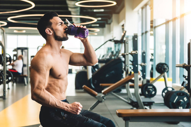 Handsome young sporty male bodybuilder drinking water while resting between exercises - Foto, Imagen