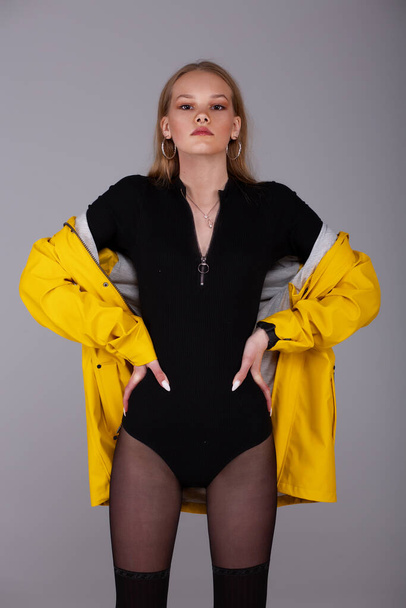 A girl in a yellow rain jacket and a black bodysuit posing in the studio. Thin waist. Hands on the belt. Youth style. Fashion. - Foto, immagini