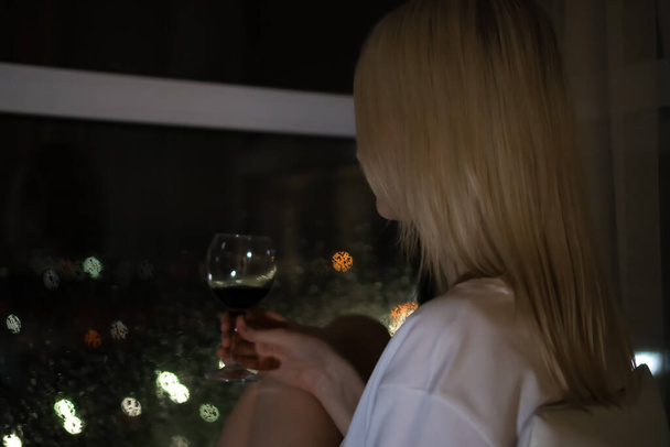Portrait of a young blonde in a white shirt with a glass of red wine looking out the window at the night of the city. Selective focus. Photo from the back - Foto, immagini