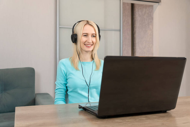 Kulaklık takan genç, gülümseyen sarışın kadın bir masada oturuyor ve dizüstü bilgisayarına bakıyor. - Fotoğraf, Görsel