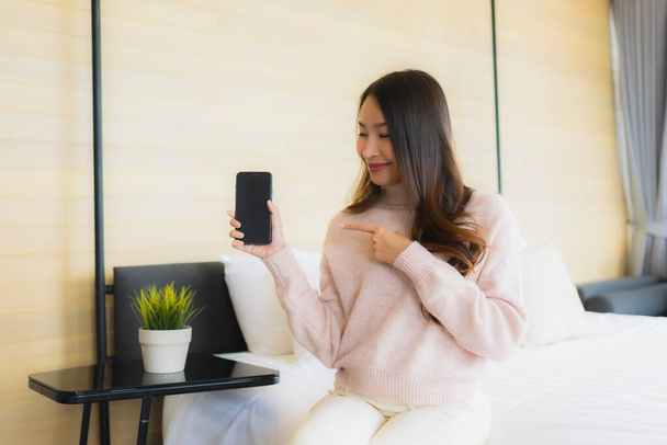 Портрет красива молода азіатка з чашкою кави і мобільним телефоном на ліжку в інтер'єрі спальні
 - Фото, зображення