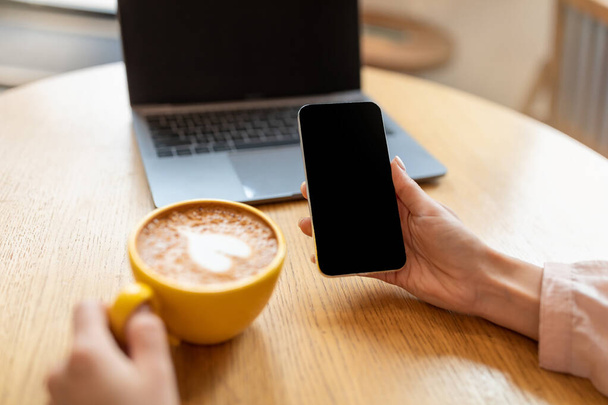 Femme méconnaissable utilisant un téléphone portable avec écran noir tout en buvant du café au café, maquette pour votre conception - Photo, image