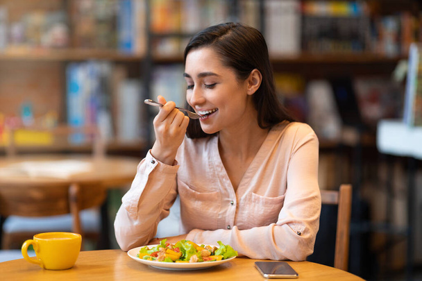 Happy latin woman eating lunch in cafe, enjoying delicious salad with closed eyes and drinking hot beverage. - Foto, imagen