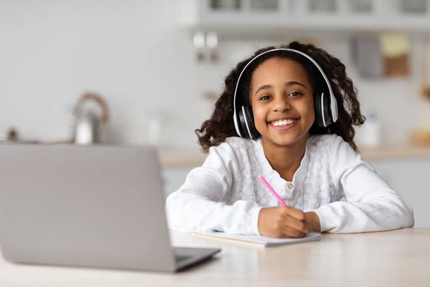 Linda chica negra con auriculares utilizando el ordenador portátil en la cocina - Foto, Imagen