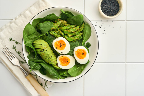 Fresh vegetable salad with avocado, asparagus, crumpled eggs with black sesame seeds and spinach on plate on light slate, stone or concrete background. Balanced lunch in bowl. Top view. Mock up. - 写真・画像