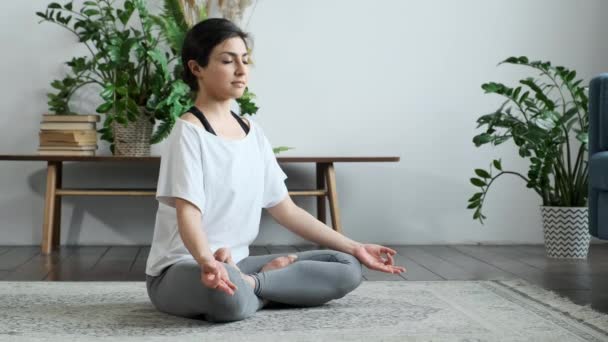 Bella giovane donna sta praticando yoga a casa rilassarsi in posizione semplice del corpo seduto sul pavimento con bei mobili intorno. Stile di vita sano e concetto di persone. - Filmati, video
