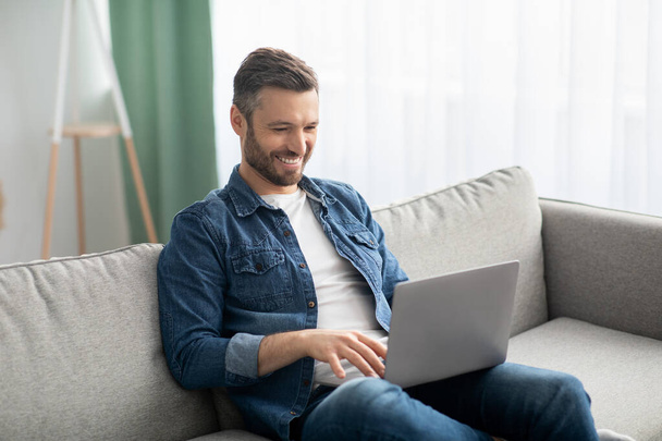Усміхнений чоловік середнього віку сидить на дивані, використовуючи ноутбук
 - Фото, зображення