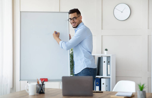 Online education. Professional Arab male tutor writing on empty blackboard with mockup, giving web lesson on laptop - Foto, imagen