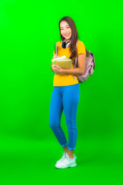 Retrato hermosa joven estudiante asiática mujer con libro teléfono auriculares y bolso en fondo verde - Foto, imagen