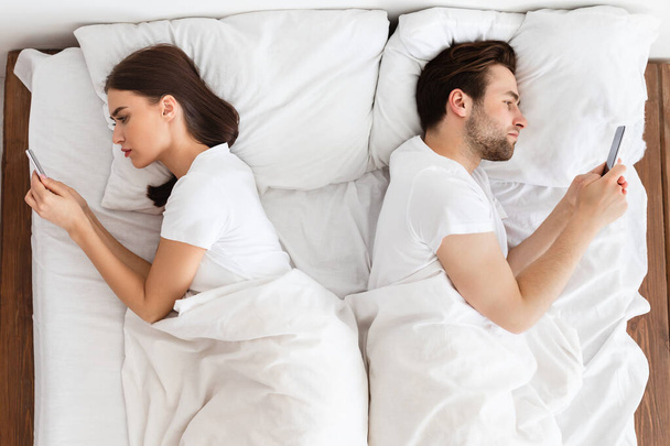 Indifferent Married Couple Texting On Phones Lying Back-To-Back Indoor, Above-View - Foto, Bild