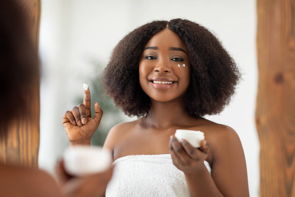 Organic cosmetics for healthy skin. Millennial black lady applying facial cream from jar in front of mirror at home - Fotó, kép