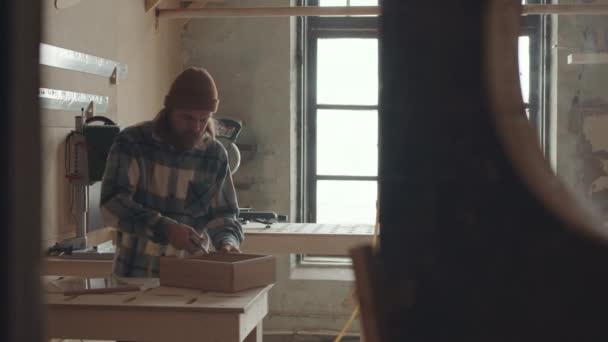 plano medio de barbudo caucásico manitas de pie en el banco de trabajo y soplado de serrín de cajón de madera hecho a mano utilizando herramienta automática - Metraje, vídeo