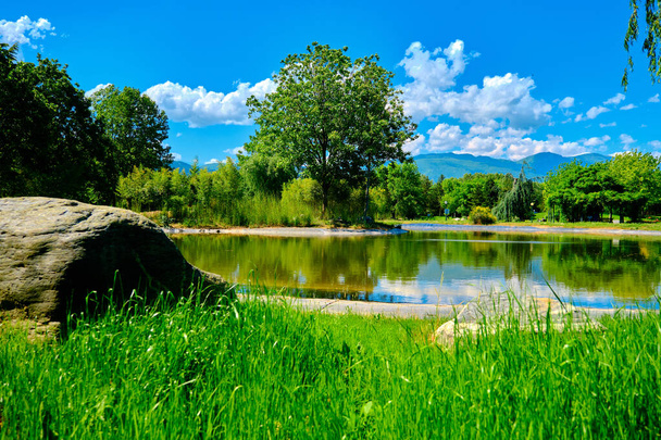 晴れた日にブルサの緑の公共および自然公園。緑の芝生の後ろに小さな池と青い空と白い雲が一日の朝と春の時間に新鮮な木がある公園. - 写真・画像