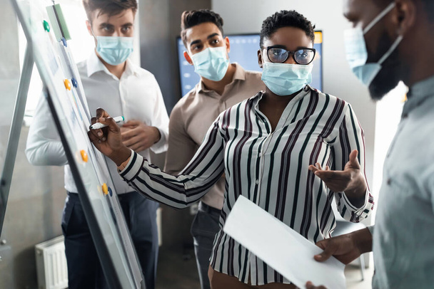 Fekete hölgy orvosi maszkban bemutatja üzleti stratégiáját a táblán - Fotó, kép
