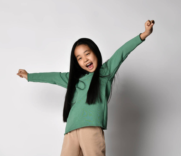 Happy screaming, singing loudly Asian kid girl in green hoodie and pants stands with arms up and outstretched - Foto, Imagen