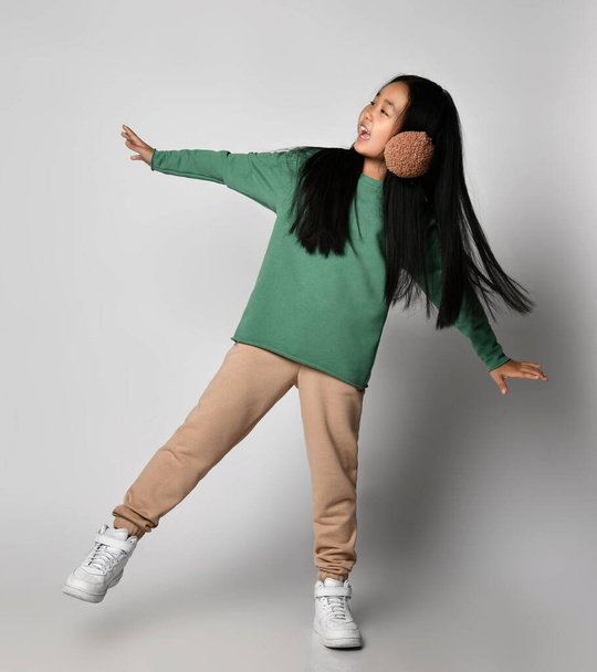 Asian kid girl in green sportswear and white sneakers stands on one leg with arms outstretched, balancing - Fotografie, Obrázek